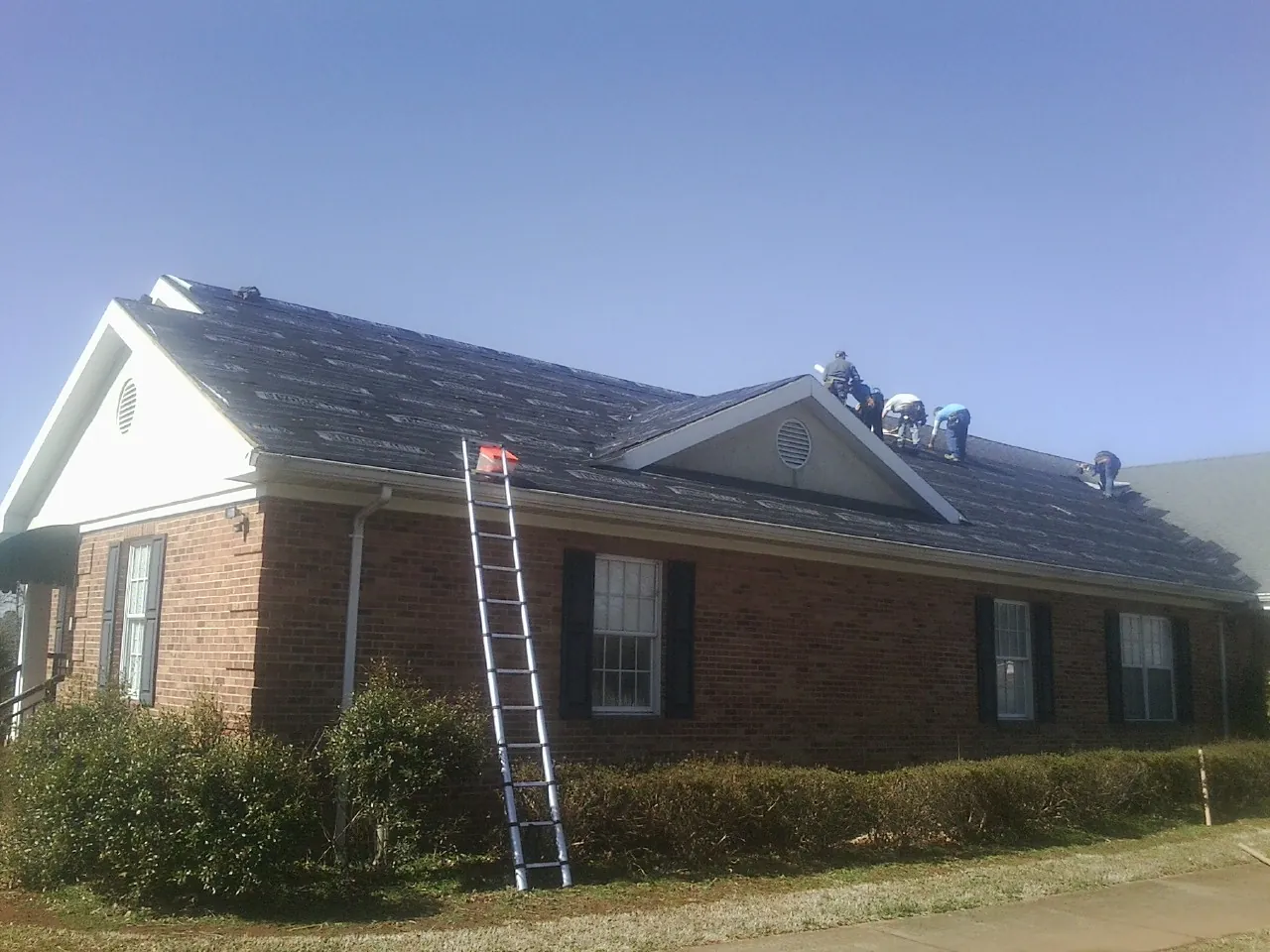 Crew tearing shingles off a brick ranch roof with a ladder set — Emergency Roof Repair work by our crew in Conway